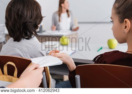 Kid Passing Message To Schoolgirl Near Teacher On Blurred Background.