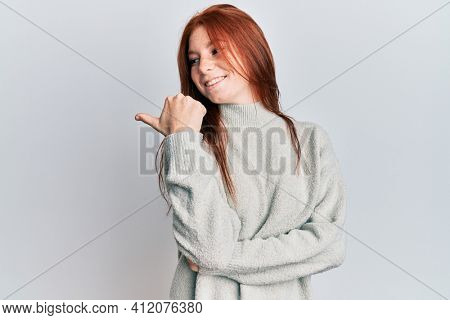 Young red head girl wearing casual winter sweater pointing thumb up to the side smiling happy with open mouth 