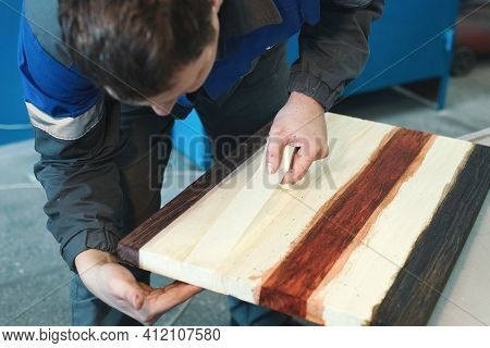 A Cabinetmaker Works With Wood In A Carpentry Shop. A Young Worker Of Caucasian Appearance In A Unif