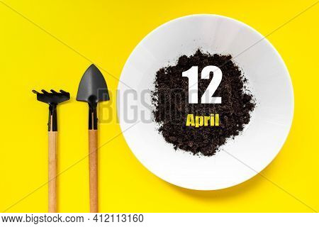 April 12nd. Day 12 Of Month, Calendar Date. White Plate Of Soil With A Small Spatula And Rake On Yel