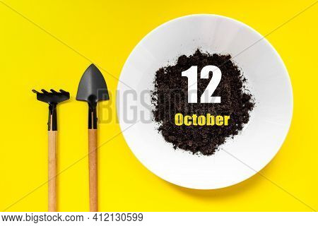 October 12nd. Day 12 Of Month, Calendar Date. White Plate Of Soil With A Small Spatula And Rake On Y
