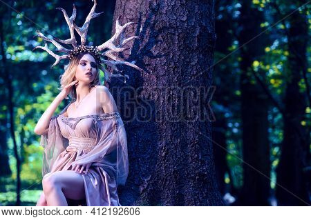 Portrait Of Beautiful Girl Posing With Artistic Deer Horns In Summer Forest Near High Tree And Weari