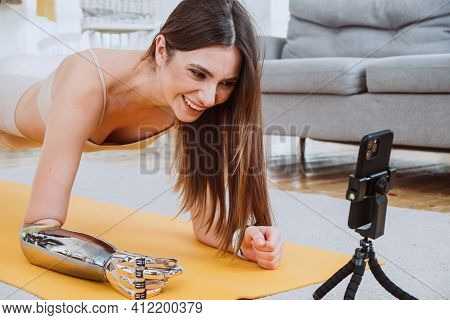 Joyful Slim Blonde Woman With Metal Prosthesis Arm Does Yoga Exercises Near Phone On Tripod In Livin