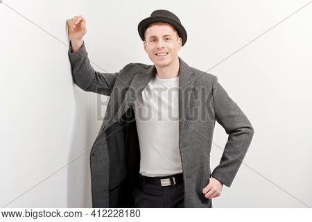 Porttrait of a handsome middle-aged man in an elegant coat and a hat standing by a white wall and smiling. Men's beauty, fashion. Lifestyle. Hipster man. 