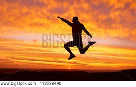 Silhouette Of A Happy Man Traveler With A Raised Hand In A Jump On A Sunset Background. Success Conc