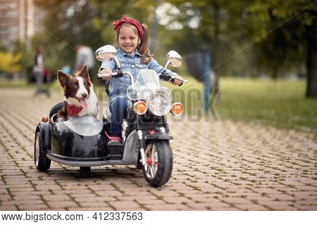 cute little girl  driving electrical motorcycle toy with sidecar and her dog in it. looking at camera, eye contact, smiling