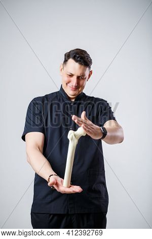 Young Male Doctor Chief Physician In A Black Surgical Suit. Close-up Portrait. Holds A Femur In His