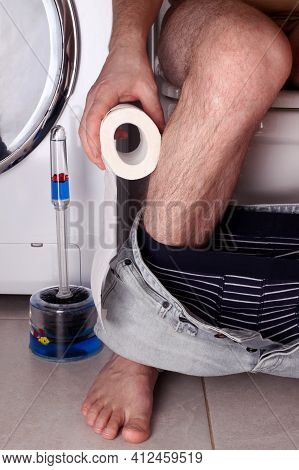 Man Sitting On Toilet Bowl At Home Or Hotel, With Toilet Paper Roll In Hands. Abdominal Stomachache,