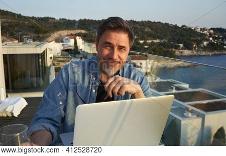 Happy man working online with laptop computer on terrace. Home office, browsing internet, tele working, video chat. Businessman on vacation at sea. Escape, freedom, getaway.