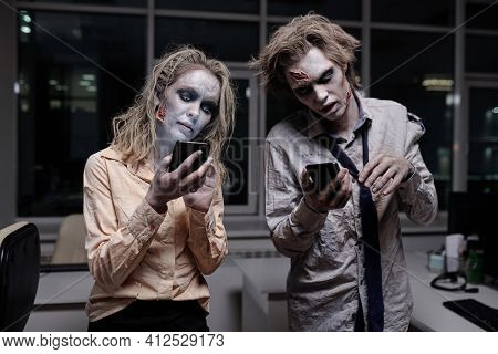 Dead and spooky businesspeople with zombie greasepaint on their faces and hands using smartphones