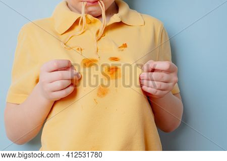Ketchup Stains On A Yellow T-shirt. Hungry Boy Licking Snack From His Fingers. Isolated On Blue Back