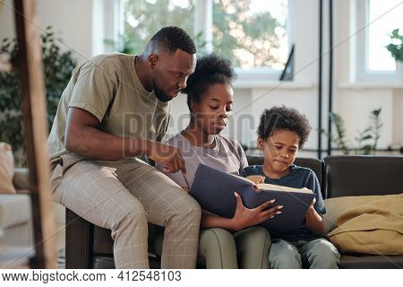 Young African family of parents and cute little son reading large book of tales or encyclopaedia