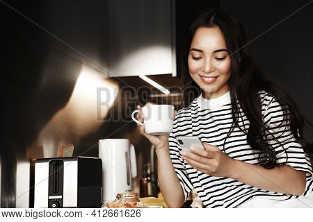 Smiling Asian Woman Reading Messages On Phone During Breakfast. Girl Drinking Coffee, Sitting In Ket