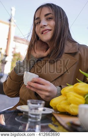 Young Beautiful Girl Drinks Espresso In A Cafe On The Street. Spring Sun In The Frame. A Bouquet Of 