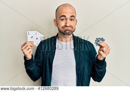 Young hispanic man playing poker holding casino cards and chips puffing cheeks with funny face. mouth inflated with air, catching air. 