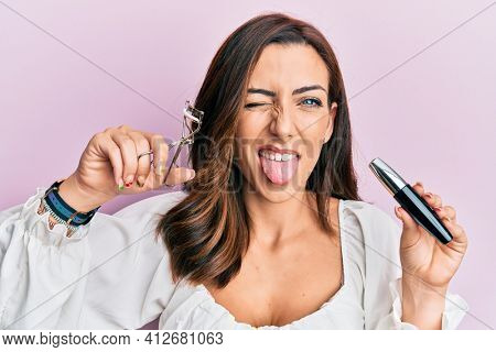 Young brunette woman holding eyelash curler sticking tongue out happy with funny expression. 