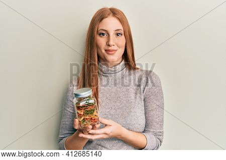 Young irish woman holding jar with farfalle pasta looking positive and happy standing and smiling with a confident smile showing teeth 