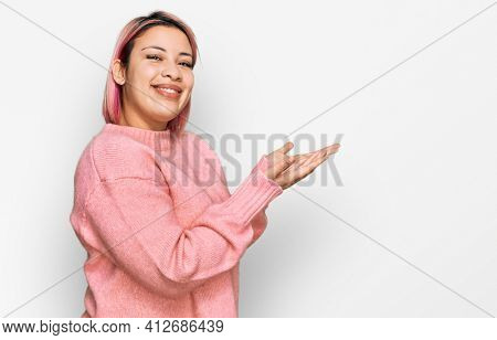 Hispanic woman with pink hair wearing casual winter sweater pointing aside with hands open palms showing copy space, presenting advertisement smiling excited happy 
