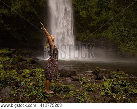 Happy Caucasian Woman Spending Time Near Waterfall, Raising Hands Up. Nature Concept. Energy Of Wate