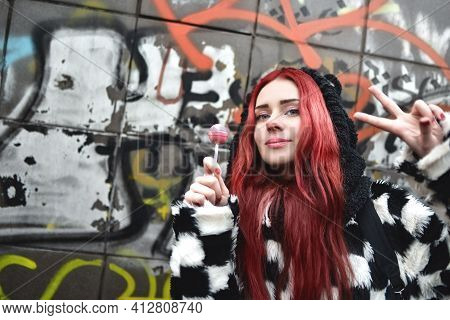 Portrait Of A Young Beauty In A Hoodie With A Delicious Pink Lollipop, Posing Against A Wall Of Graf