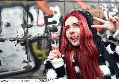 Portrait Of A Young Beauty In A Hoodie With A Delicious Pink Lollipop, Posing Against A Wall Of Graf