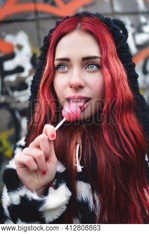 Portrait Of A Young Beauty In A Hoodie With A Delicious Pink Lollipop, Posing Against A Wall Of Graf