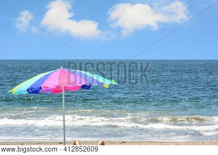 A colorful beach umbrella on the beach with a brigth blue sky an white fluffy clouds. A womans legs a just poking into the secen at the bottom of the frame.
