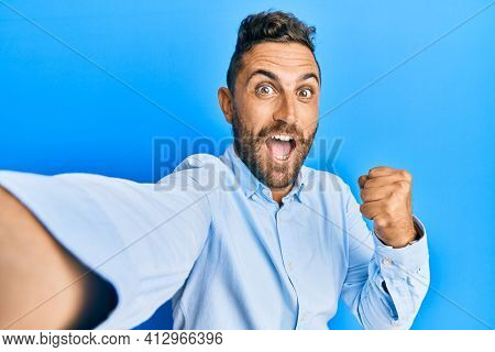 Handsome man with beard taking a selfie photo with smartphone screaming proud, celebrating victory and success very excited with raised arms 
