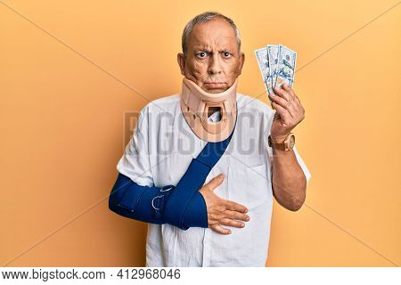 Handsome mature injured man wearing cervical collar and sling holding insurance money skeptic and nervous, frowning upset because of problem. negative person. 