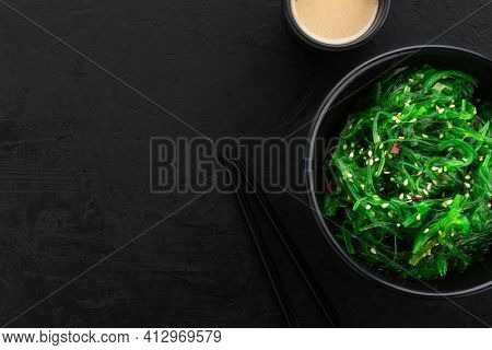 Healthy green seaweed salad with sesame seeds in a black bowl, peanut sauce and chopsticks on a slate black background. Top view, copy space.