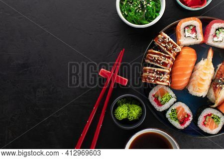Various sushi roll set served on a blue round plate, seaweed salad, soy sauce and ginger in a bowl on the side. Asian traditional food on black background, top view, copy space
