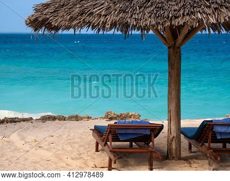 Palm Beach Umbrella And Two Empty Trestle-beds On The Turquoise Ocean Sandy Beach Under Blue Sky On 