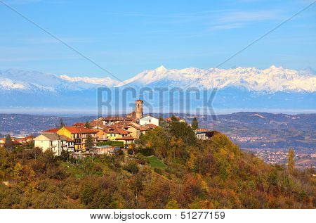 View of small town on top of the hill surrounded by autumnal trees and snowy peaks of Alps on background in Piedmont, Northern Italy.