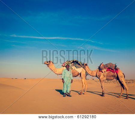 Vintage retro hipster style travel image of Rajasthan travel background - Indian cameleer (camel driver) with camels in dunes of Thar desert. Jaisalmer, Rajasthan, India
