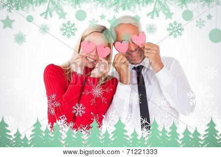 Silly couple holding hearts over their eyes against snowflakes and fir trees in green