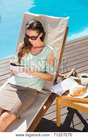 Beautiful young woman reading book by swimming pool with breakfast on table