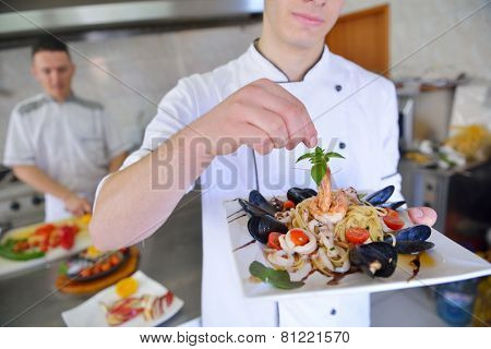 Handsome chef dressed in white uniform decorating pasta salad and seafood fish in modern kitchen