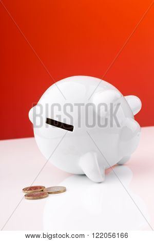White piggy bank on a red background: it's on its side with a few penny's, a concept of a tight budget. The image is tilted.