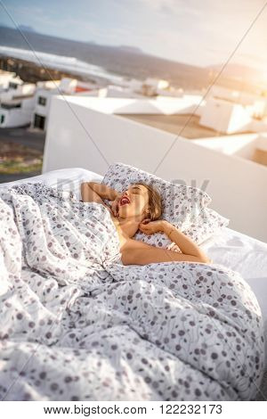 Young and cute woman straching and yawning on the bed on the roof top with white houses on the background. Good morning concept