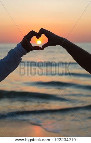 Couple doing heart shape with their hands on beachside sunset.