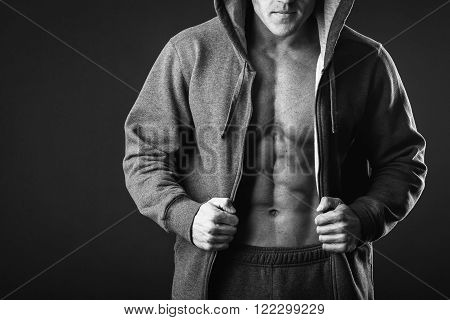 Young muscular man with open jacket revealing muscular chest and abs.Portrait of a muscular young man in hood jacket posing on black background.Young man with athletic body posing on black background