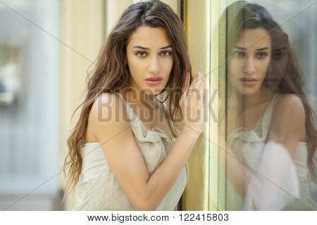 Portrait close up of young beautiful brunette woman in beige dress, Specular reflection in a shop window