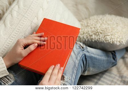 Young woman sitting on the sofa  with white cushion and holding an orange book cover on her knees, close up