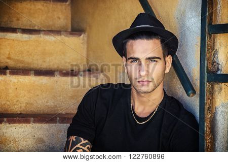 Portrait of handsome man with tattoo on hands sitting on cement stairs while leaning on wall