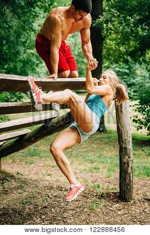 Young muscular build couple exercising on fitness trail. Male giving halping hand to woman.