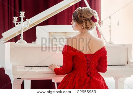 A young beautiful woman in a red evening dress sits at the grand piano