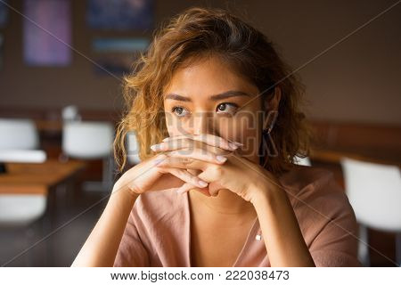 Closeup of sad young Asian woman at cafe covering nose and mouth with clasped hands and staring into vacancy. Upset manager thinking over mistakes. Failure and regret concept