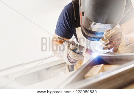 Male in face mask, protective gloves welds with argon-arc welding. Welder makes weld seam on metal frame. Worker dressed in t-shirt and overalls.