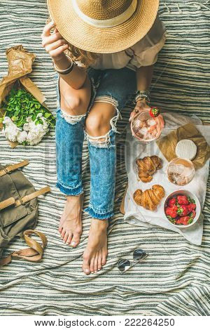 French style romantic picnic setting. Woman in shabby jeans with glass of ice rose wine, strawberries, croissants, brie cheese, hat, sunglasses, peony flowers, top view. Outdoor gathering concept