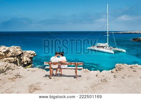 A couple sits oA couple sits on a bench and looks at the lagoon. Honeymoon lovers. Man and woman on the island. Couple in love on vacation. A voucher for a cruise trip. Sea tour. Honeymoon trip.n a bench and looks at the lagoon